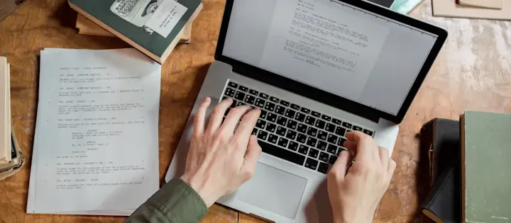 High-angle view of hands typing on a laptop surrounded by open books and writing notes